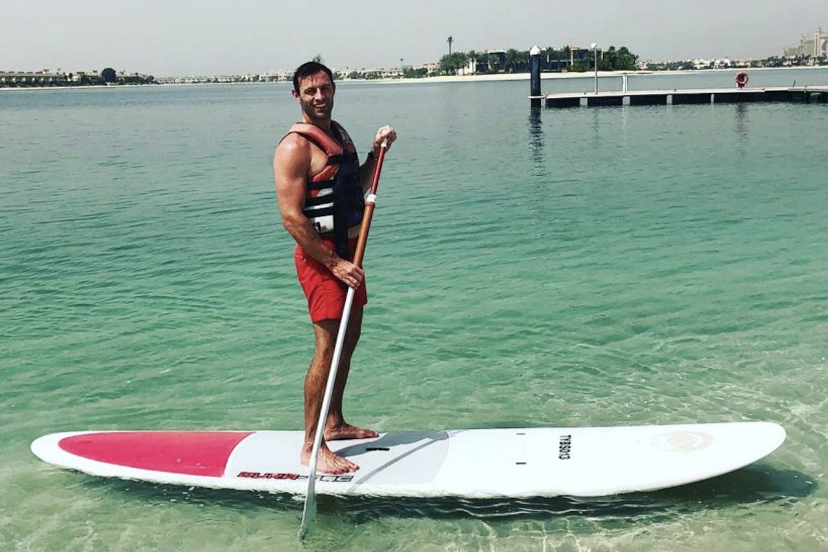 JustTwoMums top tips for dads in Dubai this summer A man in a life jacket standing on a paddleboard in clear water.