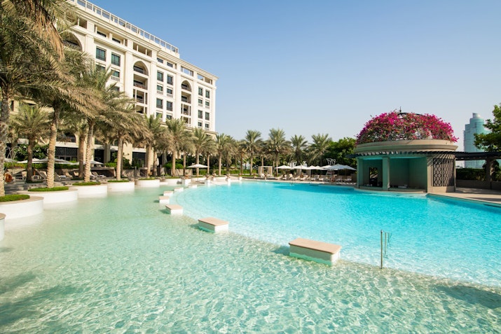 A luxurious pool with clear water, surrounded by palm trees and a hotel building in the background.
