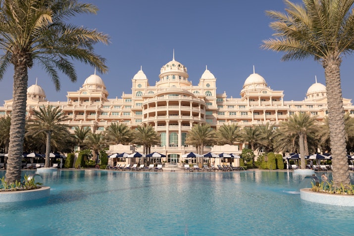 Grand hotel with ornate architecture featuring domed roofs, situated beside a large swimming pool surrounded by palm trees