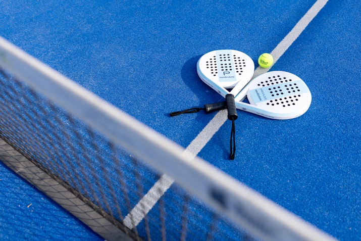 Two padel rackets and a ball on a blue court near the net.