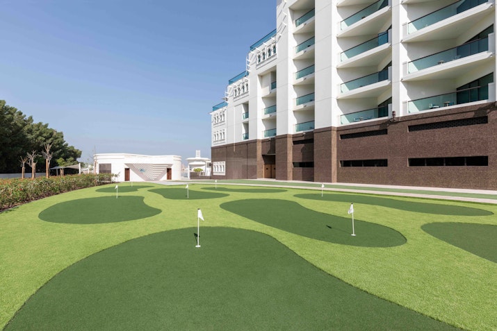 A mini golf course with a hotel building in the background, featuring well-maintained green lawns and flags at the holes.