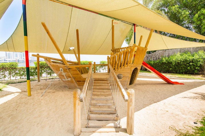 Kids’ play areas Wooden play structure resembling a ship with a slide, under a shade canopy.