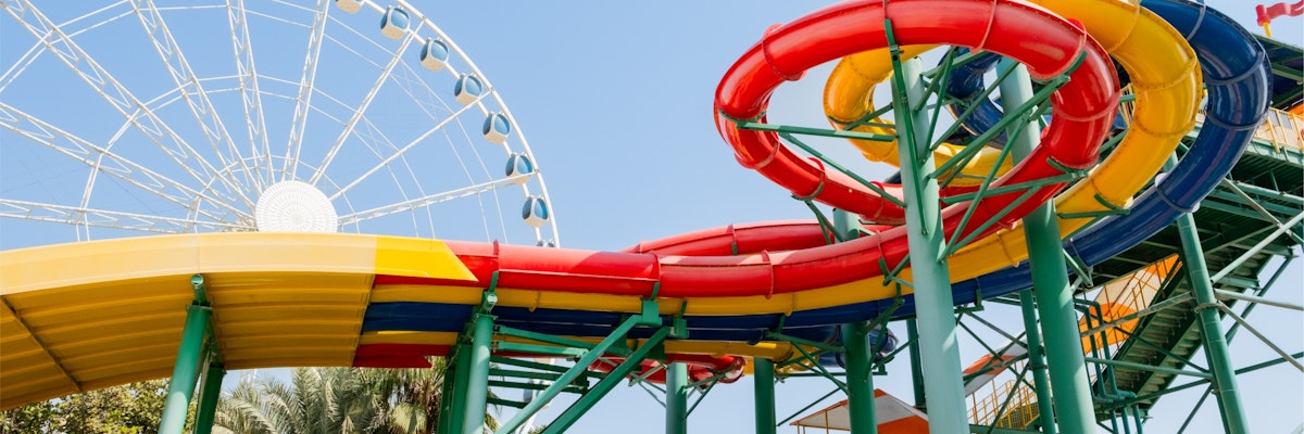 LEGOLAND® Water Park Dubai Brightly colored spiral water slides with red, yellow, and green sections at a water park in Dubai, with a large Ferris wheel in the background under a clear blue sky.