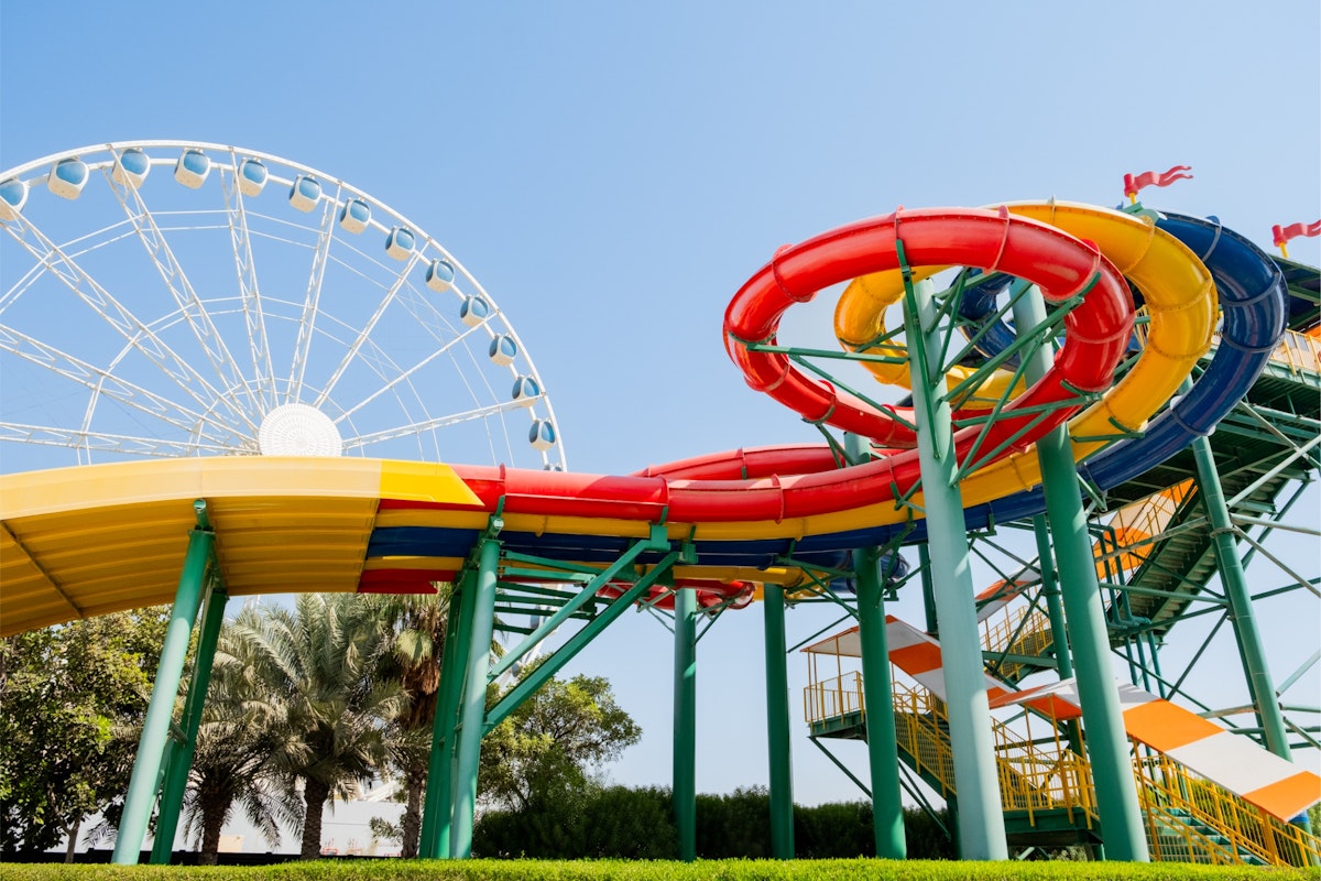 Brightly colored spiral water slides with red, yellow, and green sections at a water park in Dubai, with a large Ferris wheel in the background under a clear blue sky.
