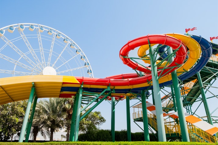 Brightly colored spiral water slides with red, yellow, and green sections at a water park in Dubai, with a large Ferris wheel in the background under a clear blue sky.