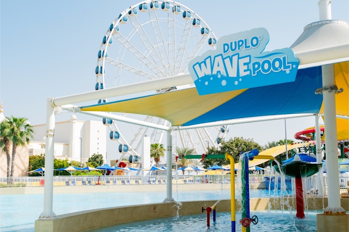 DUPLO Wave Pool area at a water park, with a large ferris wheel in the background and colorful water play structures.