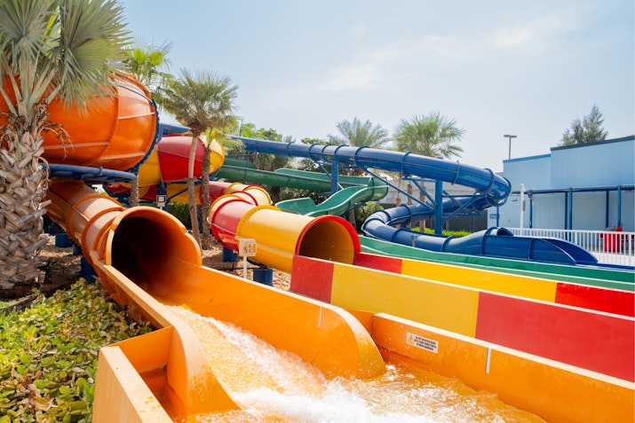 A collection of colorful water slides in shades of orange, red, green, and blue with twisting tubes and open chutes, surrounded by palm trees and tropical landscaping at a water park