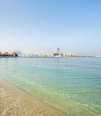 Anantara The Palm Dubai Resort A peaceful beach with clear water, surrounded by resorts and distant city skyline.