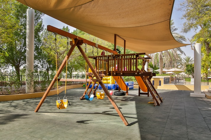 A children's playground with swings and a slide, shaded by a canopy and surrounded by trees.