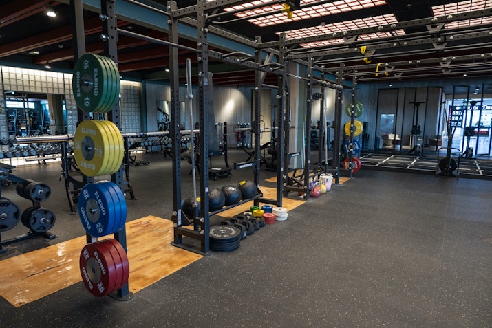 Gym weightlifting area with a variety of weight plates, squat racks, and barbells organized on rubberized flooring.