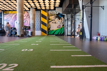 Gym area with a green turf track, pop art mural on the wall, and kettlebells lined up in a modern, well-lit setting