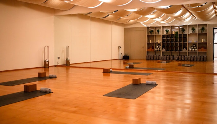 Vinyasa at 700 Padel & Fitness Hub A calming yoga studio featuring neatly arranged mats, blocks, and towels on a polished wooden floor, with soft ceiling drapes and warm lighting creating a peaceful ambiance.