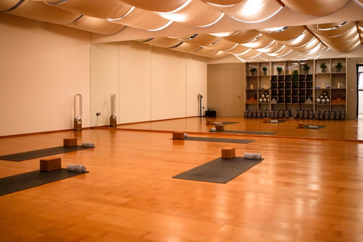 A calming yoga studio featuring neatly arranged mats, blocks, and towels on a polished wooden floor, with soft ceiling drapes and warm lighting creating a peaceful ambiance.