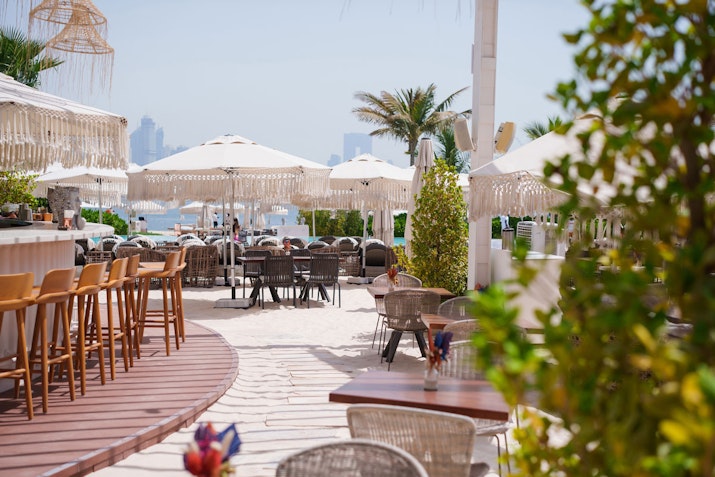 Beachside outdoor dining area with umbrellas, bar stools, and woven chairs.