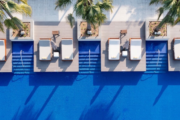 An aerial view of a luxurious pool area with sun loungers and palm trees, showcasing blue water and shaded seating.