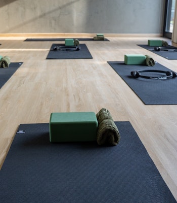 Mantra Yoga and Pilates A serene yoga studio with neatly arranged black mats, each equipped with a green yoga block, a rolled towel, and a Pilates ring, creating a welcoming space for yoga or Pilates practice. The studio features light wooden flooring, soft natural lighting from large windows, and minimalist decor, fostering a calm and focused environment for wellness activities