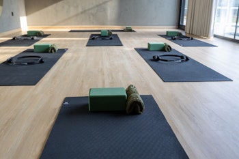 A serene yoga studio with neatly arranged black mats, each equipped with a green yoga block, a rolled towel, and a Pilates ring, creating a welcoming space for yoga or Pilates practice. The studio features light wooden flooring, soft natural lighting from large windows, and minimalist decor, fostering a calm and focused environment for wellness activities