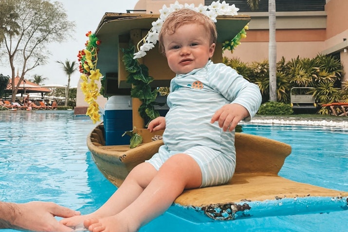 A baby sitting in a small boat in a pool, wearing a striped onesie