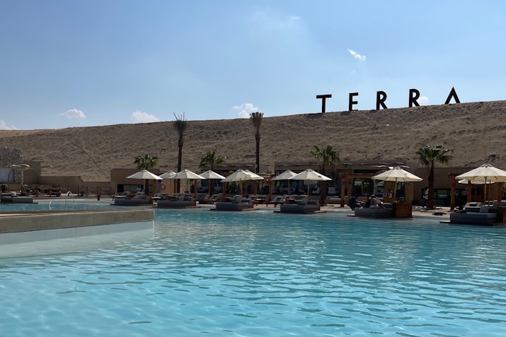 A pool with umbrellas and seating, set against a large "TERRA" sign on a sandy hill.