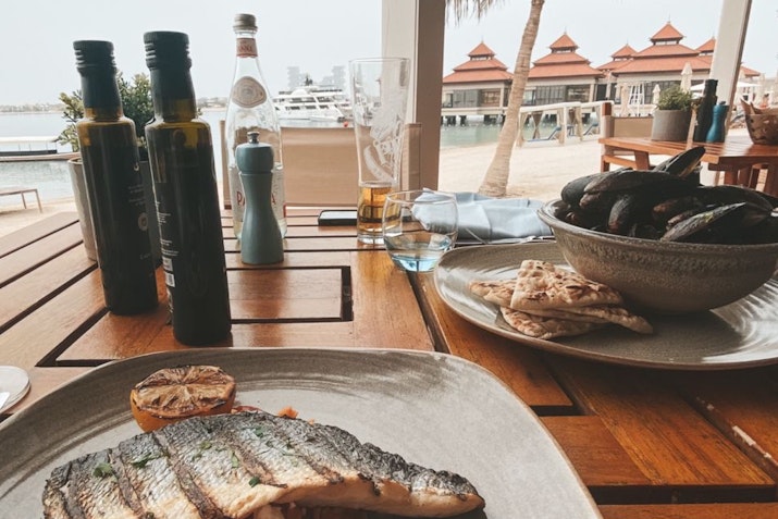A table set with grilled fish, a bowl of mussels, and drinks with a scenic view of the waterfront.