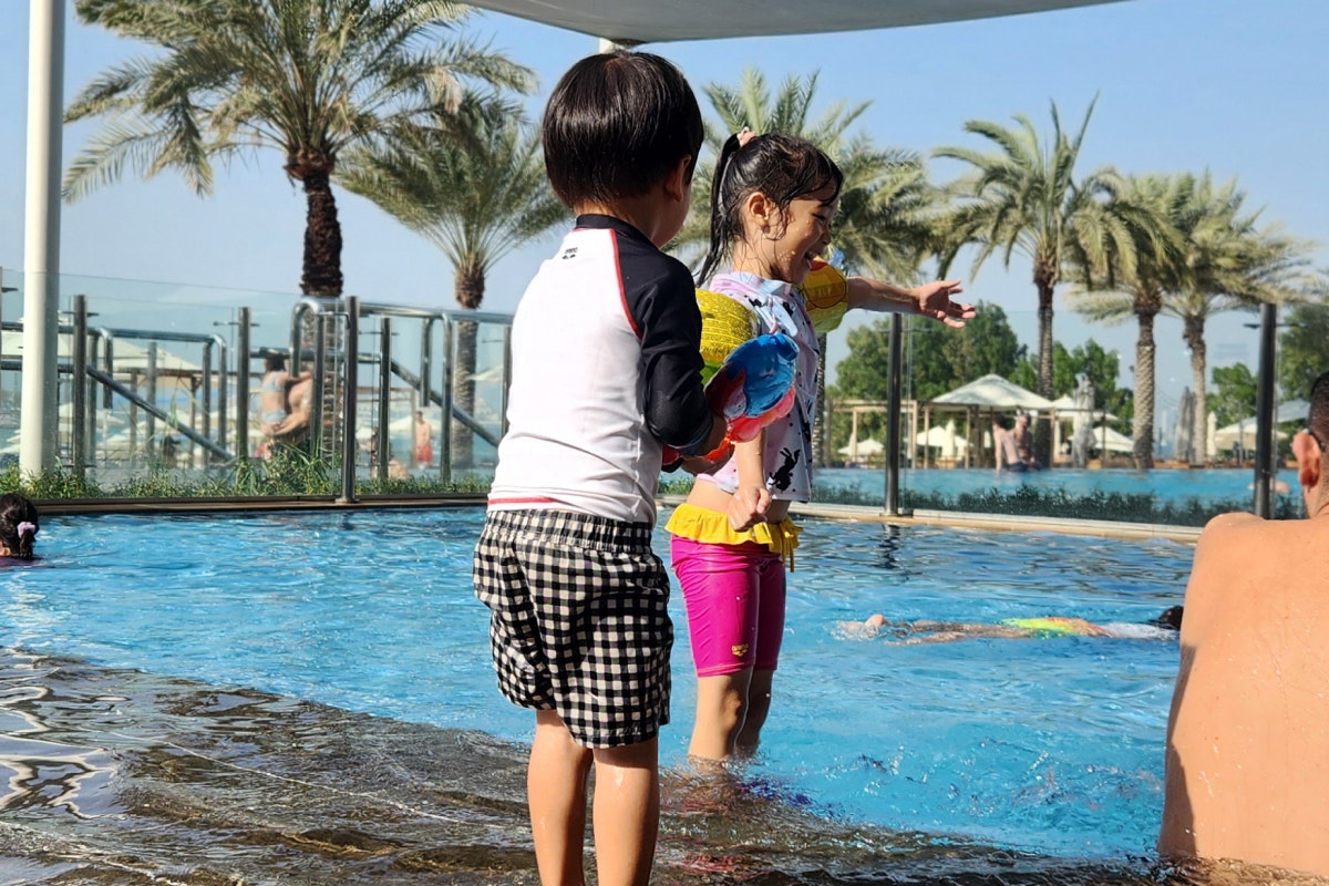 Two children playing in the pool, with one holding a water gun, and palm trees in the background.