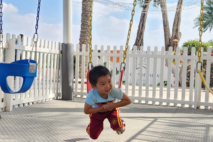 A young boy playing on a swing set with a fence and palm trees in the background.