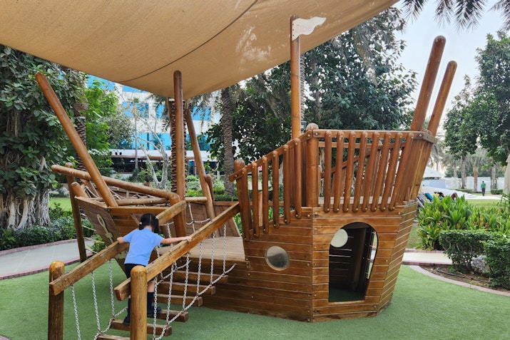 A child climbing the rope ladder on a wooden pirate ship play structure in a playground.