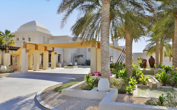 A beautifully landscaped entrance featuring palm trees, decorative pots, and a shaded walkway leading to an elegant building.
