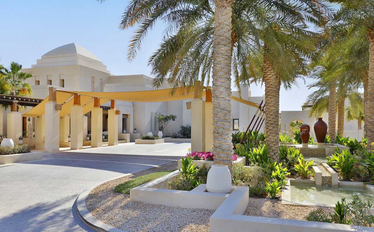 A beautifully landscaped entrance featuring palm trees, decorative pots, and a shaded walkway leading to an elegant building.