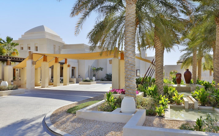 A beautifully landscaped entrance featuring palm trees, decorative pots, and a shaded walkway leading to an elegant building.