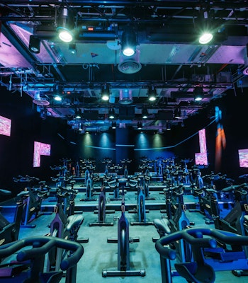 1Rebel A spacious, neon-lit indoor cycling studio with multiple rows of stationary bikes, featuring colorful LED lights and digital screens on the walls for an energizing workout atmosphere.