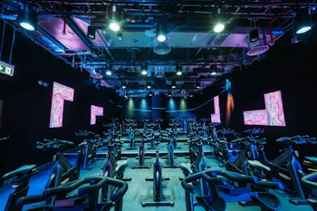 A spacious, neon-lit indoor cycling studio with multiple rows of stationary bikes, featuring colorful LED lights and digital screens on the walls for an energizing workout atmosphere.