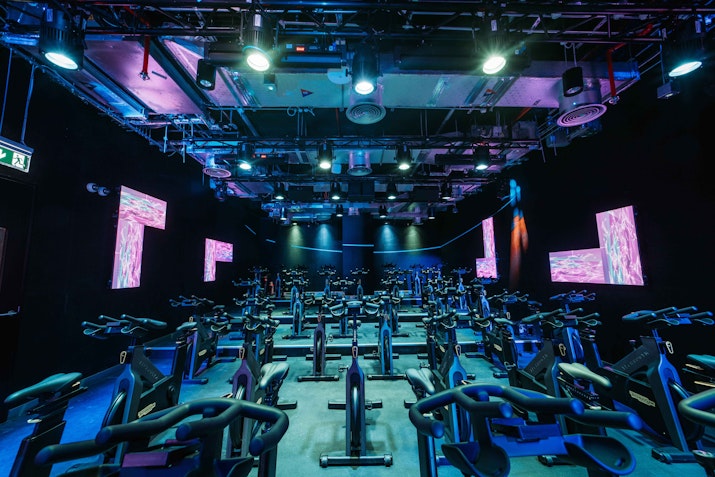A spacious, neon-lit indoor cycling studio with multiple rows of stationary bikes, featuring colorful LED lights and digital screens on the walls for an energizing workout atmosphere.
