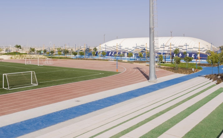 A well-maintained running track with blue and red lanes next to a green field and stadium seating, set against a bright, clear sky.