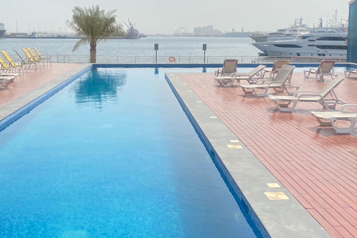 Outdoor pool area with sun loungers on a wooden deck, offering views of the harbor and docked yachts in the background.