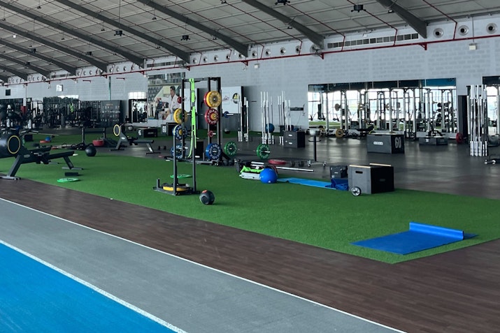Large gym featuring a turf area for functional exercises, surrounded by various workout equipment and open spaces under a high ceiling.
