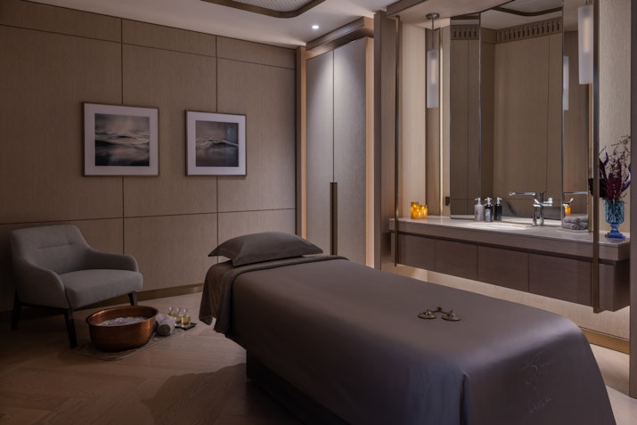A serene spa room featuring a massage bed with soft gray linens, ambient lighting, and a comfortable armchair beside a wooden bowl with towels. The modern decor includes a vanity area with a mirror and calming artwork on the walls, creating a peaceful ambiance