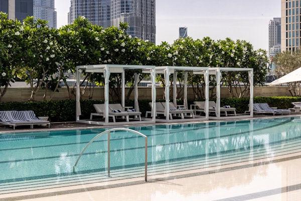 A swimming pool with lounge cabanas surrounded by greenery, set against a backdrop of city buildings.