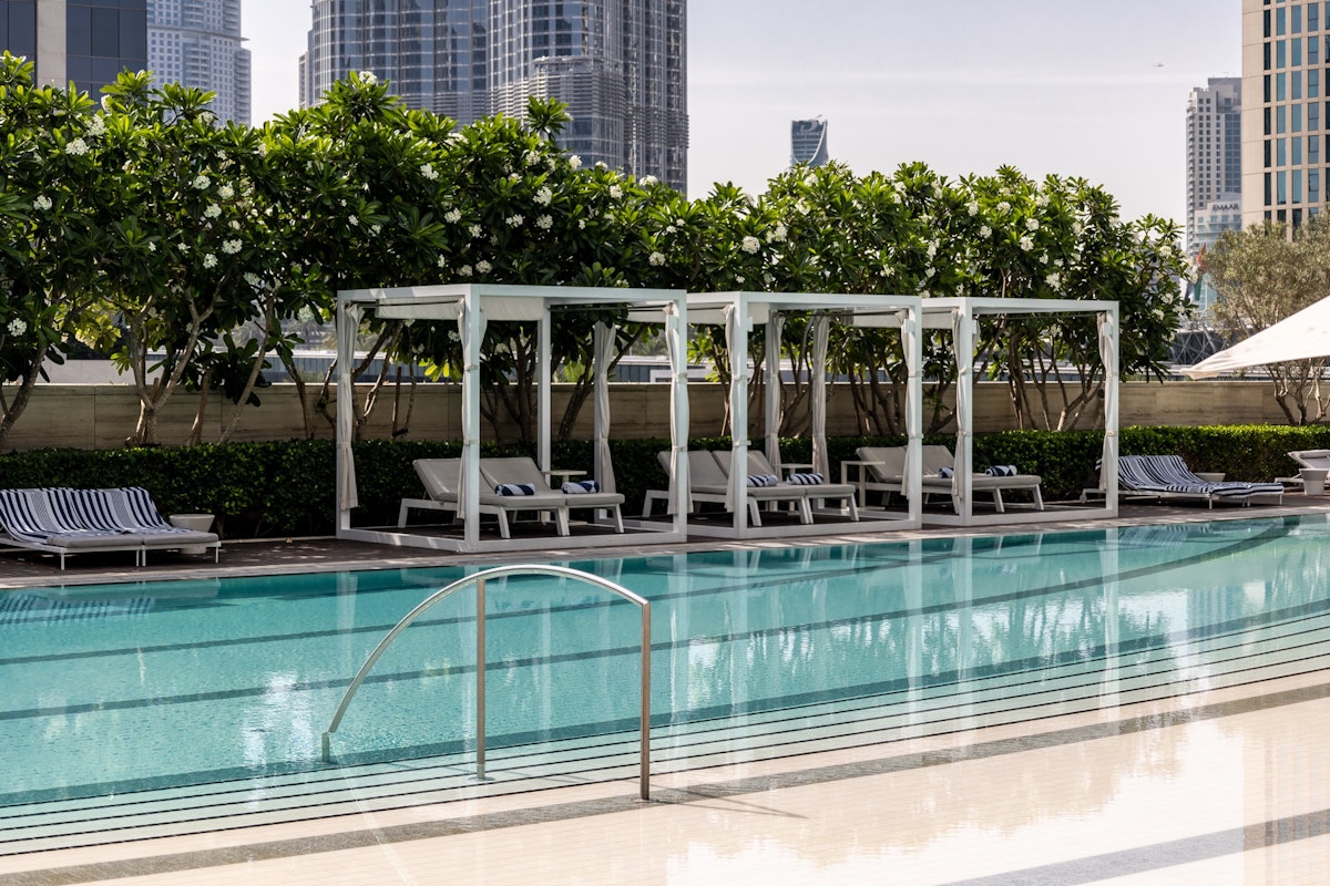 A swimming pool with lounge cabanas surrounded by greenery, set against a backdrop of city buildings.
