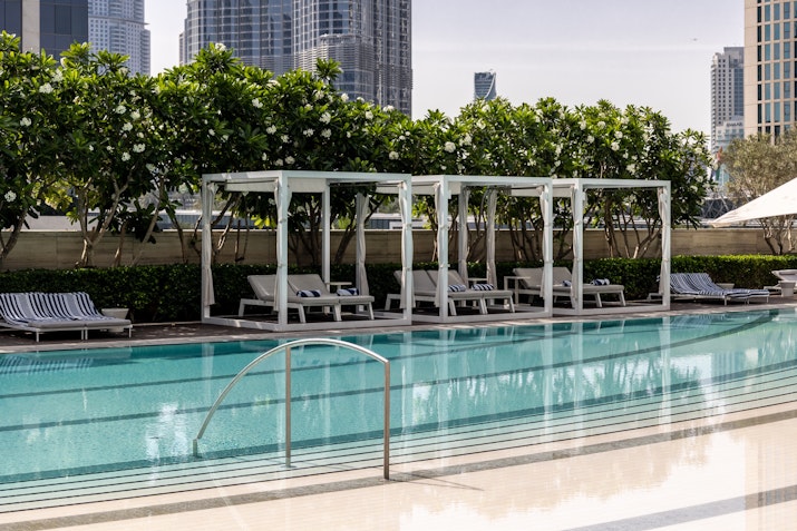 A swimming pool with lounge cabanas surrounded by greenery, set against a backdrop of city buildings.