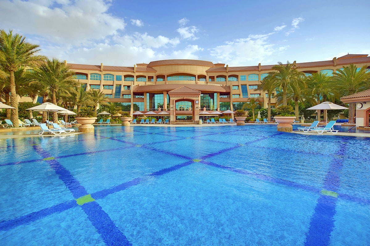 Al Raha Beach Hotel A luxurious hotel with a large swimming pool in front, surrounded by palm trees and sun loungers under umbrellas, set against a partly cloudy sky.