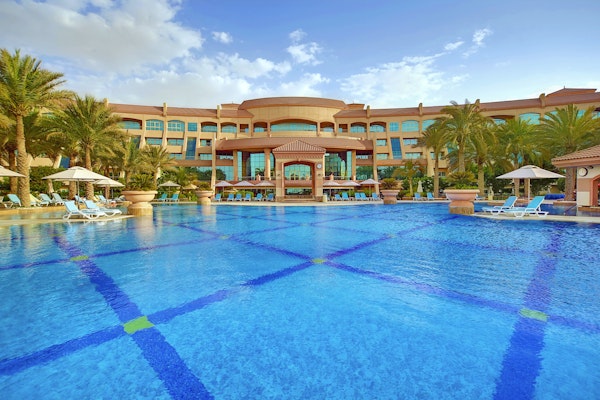 A luxurious hotel with a large swimming pool in front, surrounded by palm trees and sun loungers under umbrellas, set against a partly cloudy sky.