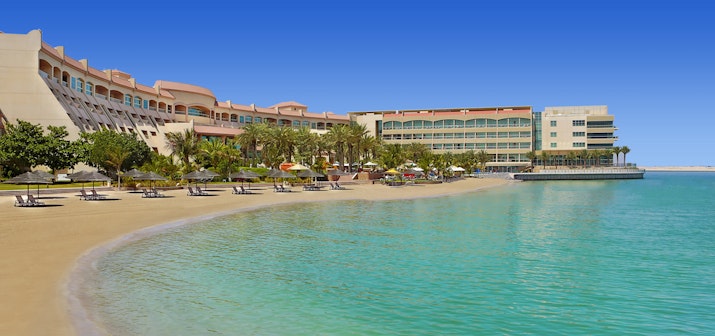 Al Raha Beach Resort & Spa A beachfront hotel with a large outdoor area and sun loungers, overlooking clear turquoise waters and a bright blue sky.