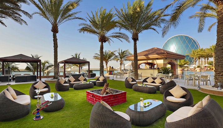 An outdoor lounge area with comfortable seating, surrounded by palm trees and a fire pit, featuring a bar structure and a modern dome-shaped building in the background.