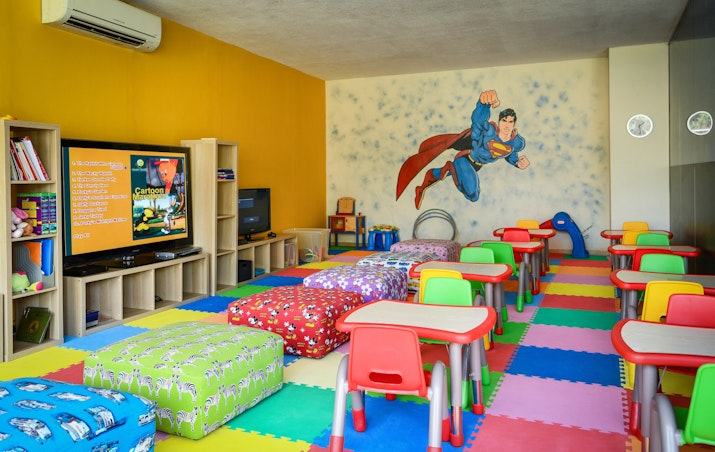 A colorful children's playroom with small tables, cushioned seating, and a mural of a superhero on the wall, featuring a television and various toys.