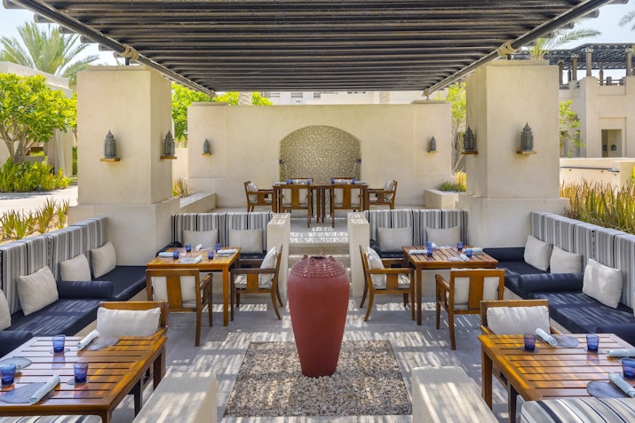 An outdoor dining area with wooden tables, cushioned seating, and a decorative pot at the center, surrounded by lush greenery and a serene atmosphere.