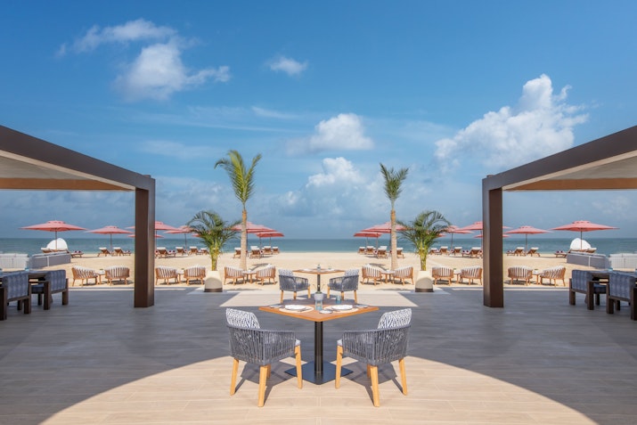 Outdoor dining area with modern chairs and tables, overlooking a beach with umbrellas and palm trees under a bright blue sky.
