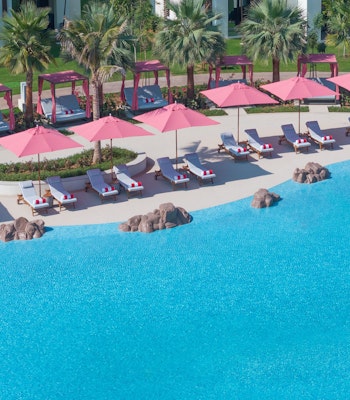 Anantara Mina Al Arab Ras Al Khaimah Resort Aerial view of a poolside area with pink umbrellas and lounge chairs surrounded by palm trees.