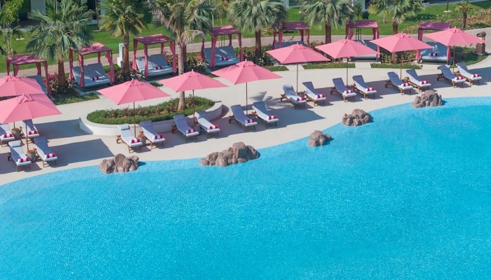 Anantara Mina Al Arab Ras Al Khaimah Resort Aerial view of a poolside area with pink umbrellas and lounge chairs surrounded by palm trees.