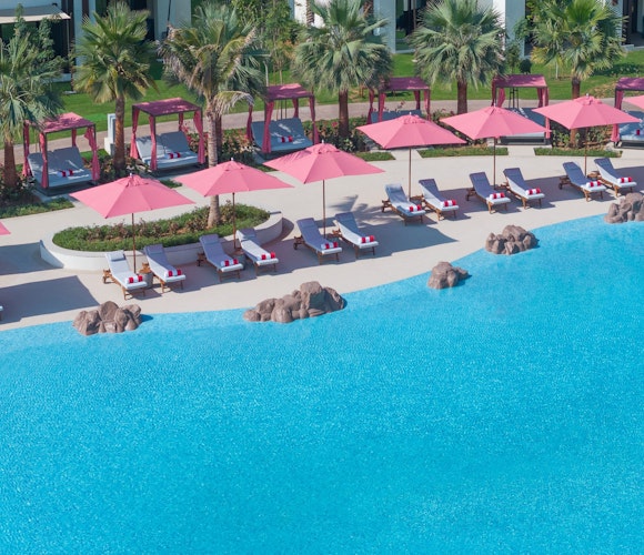 Anantara Mina Al Arab Ras Al Khaimah Resort Aerial view of a poolside area with pink umbrellas and lounge chairs surrounded by palm trees.